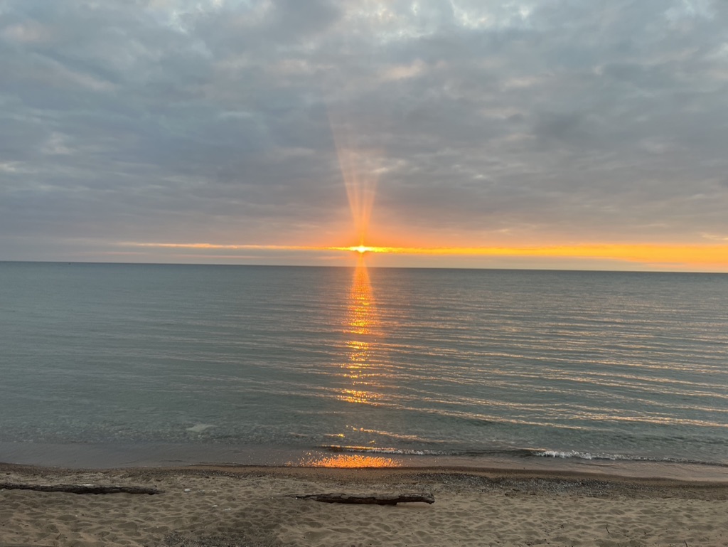 Golden sunlight breaks through clouds at the horizon and reflects across calm water towards a sandy shore.