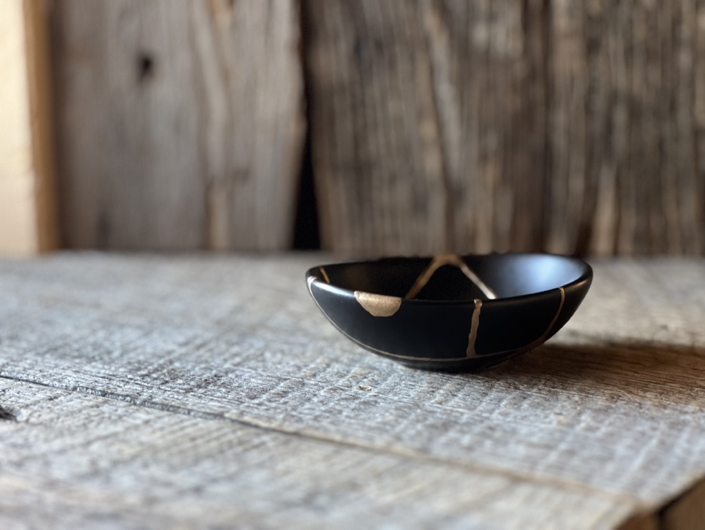 A small black dish with golden repair seams resting on a weathered wooden table.