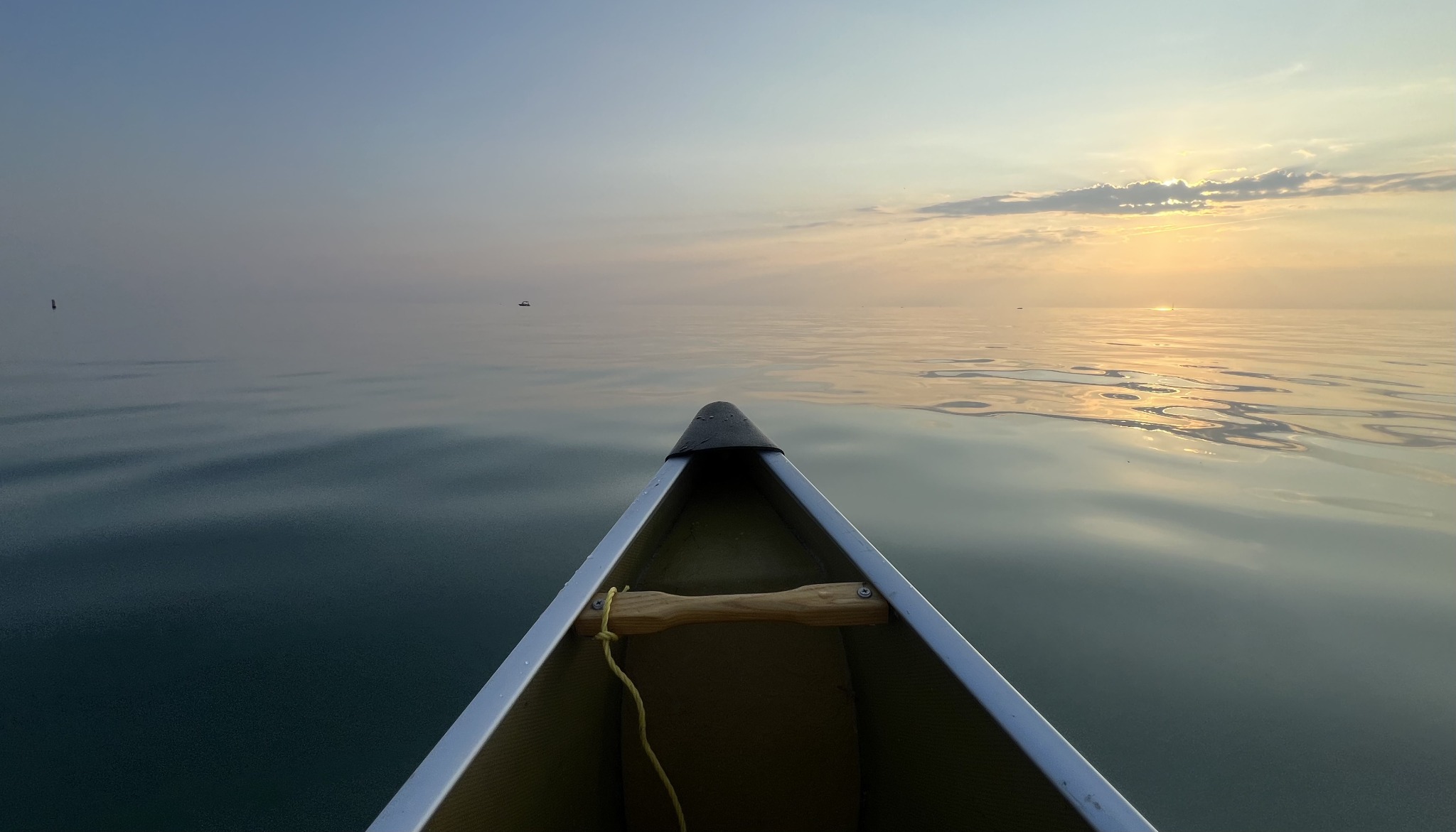 The bow of a canoe pointing across calm water toward a soft golden sunset.