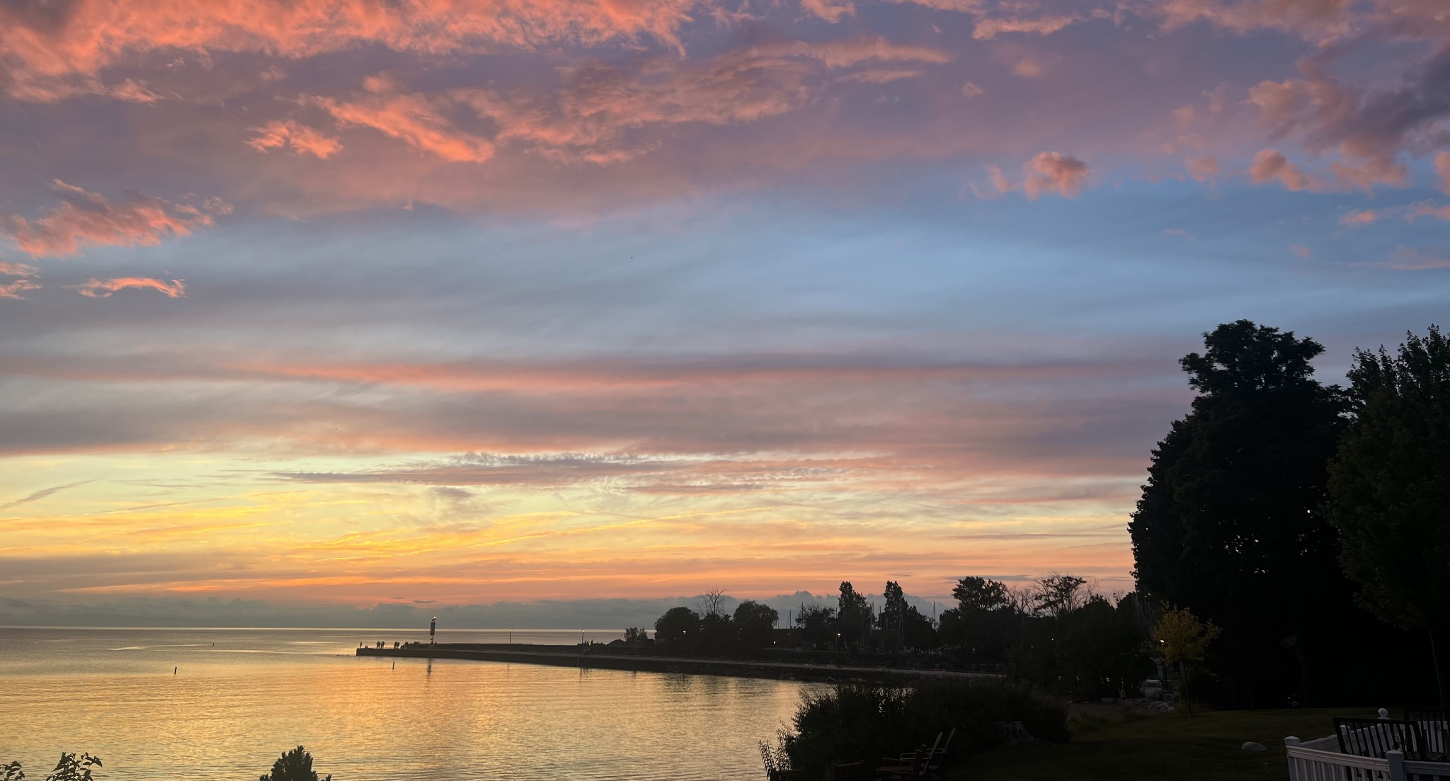 Pink and orange clouds over a lake and pier at sunset, with trees silhouetted along the shoreline, in a wide crop.