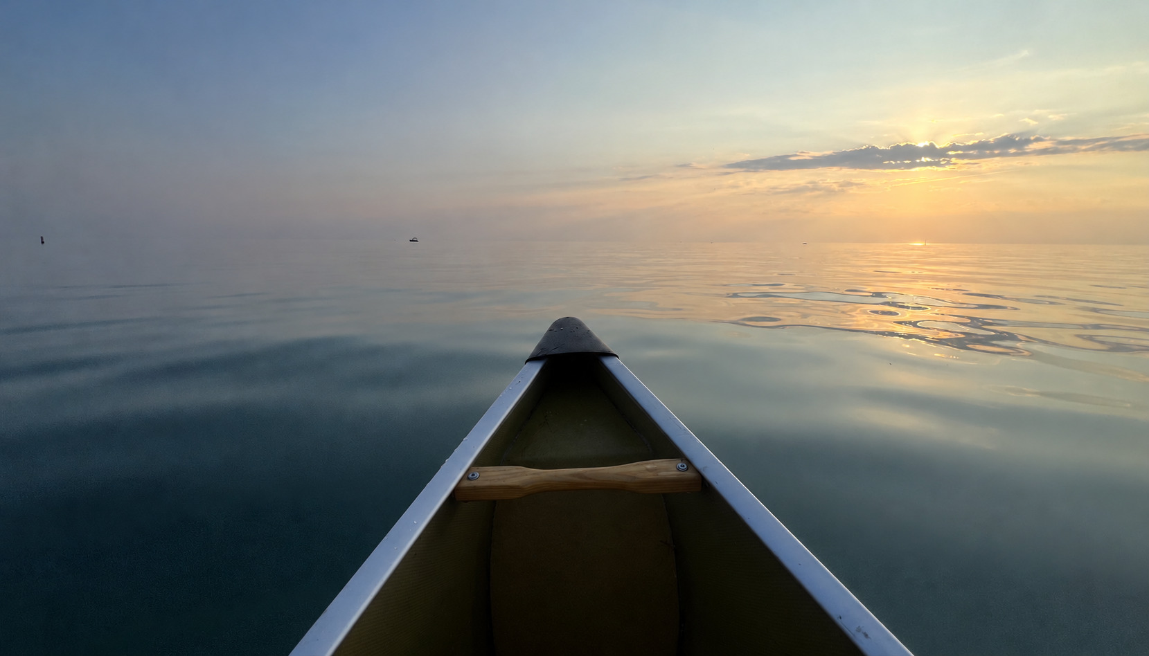 The bow of a canoe pointing across calm water toward a soft golden sunset.