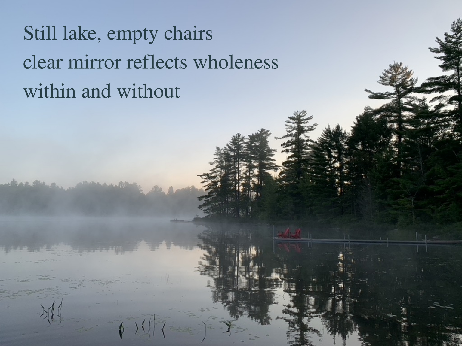 Two red chairs beside a misty lake, with pine trees and the sky reflected in still water. Haiku: Still lake, empty chairs / clear mirror reflects wholeness / within and without